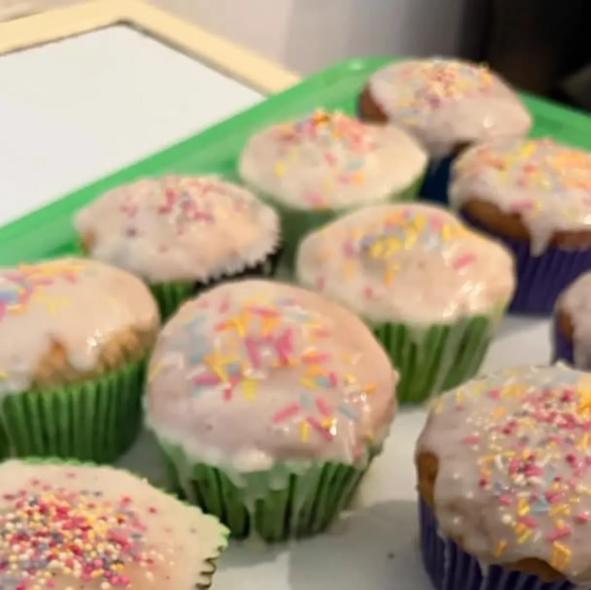 A tray of freshly iced muffins in green and purple cases, topped with pastel sprinkles.